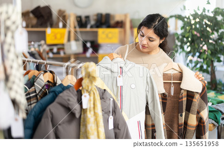 Latin girl is busy looking at clothes items in store, she chooses winter coat or jacket Latin girl is busy looking at clothes items in store, she chooses winter coat or jacket 135651958