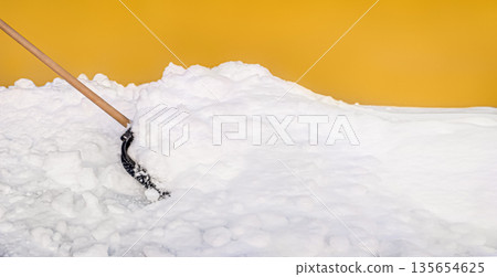 Snow shovel scooping fresh snow against a vibrant yellow wall 135654625