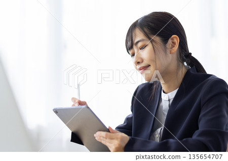 A young woman in a suit operating a tablet at a desk in a bright room A young woman in a suit operating a tablet at a desk in a bright room 135654707