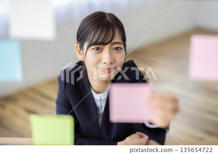 A young woman in a suit smiling with a sticky note in her hand in the office 135654722