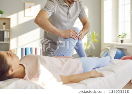 Male physiotherapist examining a young woman's knee on the couch in rehabilitation clinic. 135655318