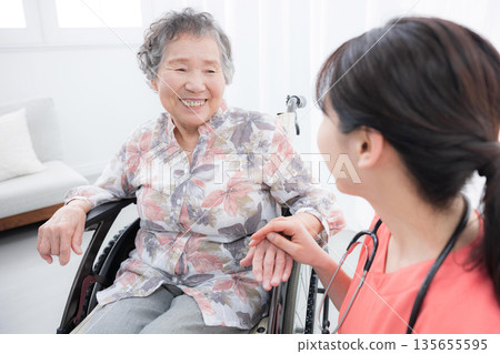 Image of a nurse supporting an elderly person in a wheelchair in a hospital or visiting care facility, holding their hand and looking at each other 135655595