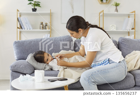 Mother measuring temperature of ill child on sofa, administering care and comfort 135656237