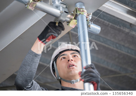 施工工作,例如用夾具將單根管道固定在靠近天花板的高腳手架上。圖片中可以看到建築工人和工人。扳手。 施工工作,例如用夾具將單根管道固定在靠近天花板的高腳手架上。圖片中可以看到建築工人和工人。扳手。 135656340