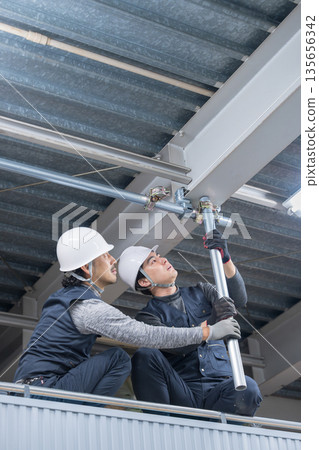 Construction work such as clamping single pipes on high scaffolding near the ceiling. Image of construction workers and workers tightening with wrenches. Construction work such as clamping single pipes on high scaffolding near the ceiling. Image of construction workers and workers tightening with wrenches. 135656342