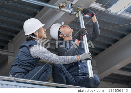 Construction work such as clamping single pipes on high scaffolding near the ceiling. Image of construction workers and workers tightening with wrenches. 135656343