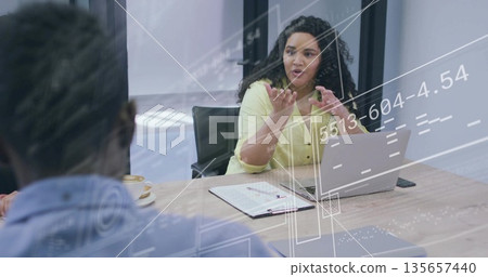 Gesturing woman wearing yellow blouse speaking at conference room table, with laptop and clipboard 135657440