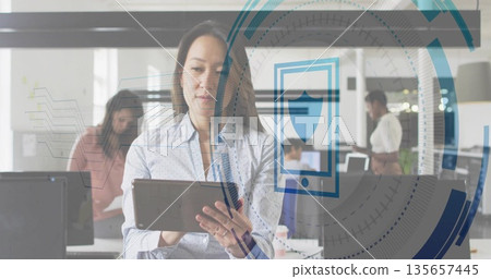 Interacting mid-adult woman holding tablet in open-plan office, with colleagues reviewing documents 135657445