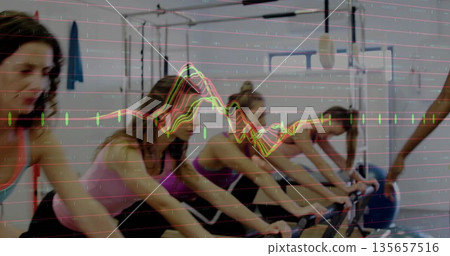 Pedaling six women in athletic wear on bikes in gym, with microphone boom, blue exercise ball 135657516