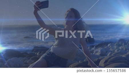 Woman in knitted top and denim shorts taking selfie on coastal rocks at dusk holding smartphone 135657586