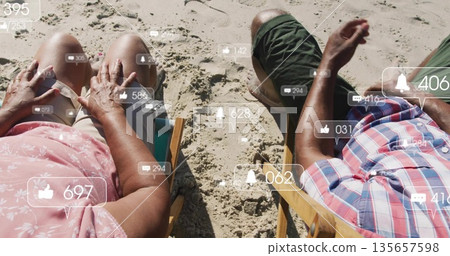 Sitting senior couple viewing floating social media icons on wooden deck chairs on sandy beach 135657598