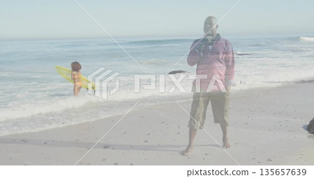 Sipping from cup senior man wearing red shirt on sandy shoreline, with yellow surfboard, copy space 135657639