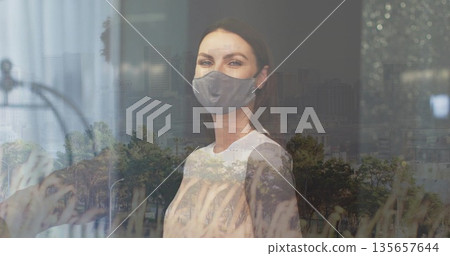 Standing woman wearing grey cloth mask and white top at window, reflecting grass and skyline 135657644