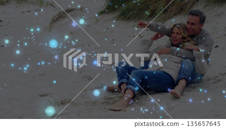 Sitting couple wearing pullover and jeans hugging on sandy beach at dune grass, glowing light Sitting couple wearing pullover and jeans hugging on sandy beach at dune grass, glowing light 135657645