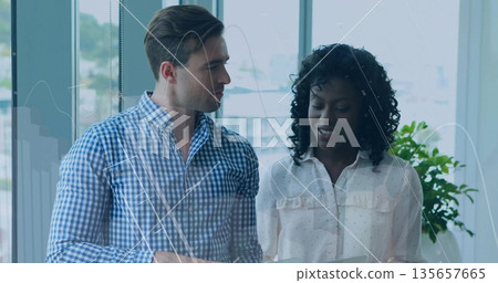 Coworkers in business attire reviewing tablet data at office windows, with plant 135657665