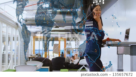 Asian woman talking on mobile phone at standing desk in office, with digital map, copy space 135657672