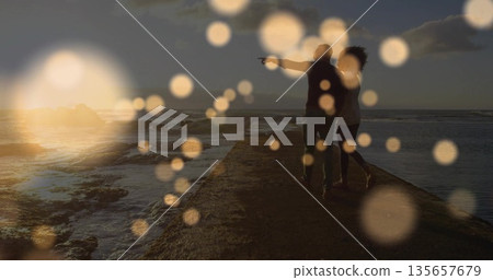 Walking couple pointing toward ocean rocks at sunset with bokeh orbs on stone pier, copy space 135657679