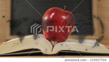 Balancing red apple on open book on wooden desk by chalkboard, with dotted world map overlay 135657704