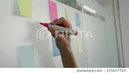 Holding hand gripping pink marker poised on glass board in office with pastel notes, copy space 135657705