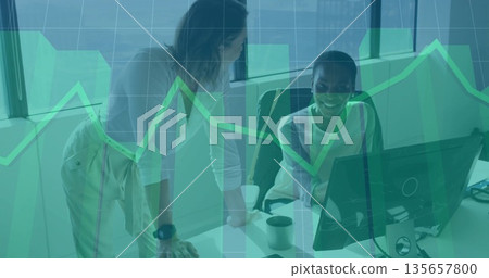 Leaning women in business casual reviewing charts at office desk, with PC monitor and smartphone 135657800