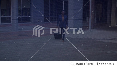 Walking business traveler pulling wheeled suitcase at plaza, with concrete columns, glass windows 135657805