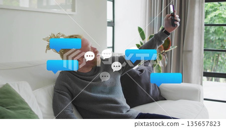 Man wearing grey sweater reclining on white sofa in living room and holding smartphone, copy space 135657823