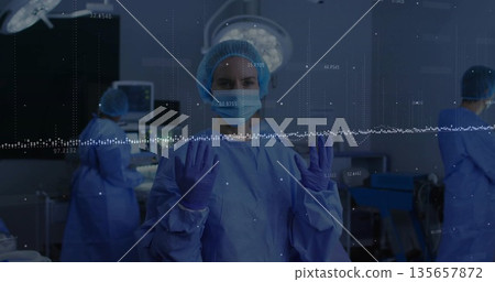Surgeon adjusting sterile gloves in operating room beneath overhead light, with waveform overlay 135657872