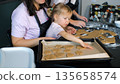 Young mother and her daughters cut ginger cookie dough and place the shaped pieces on a baking tray 135658574