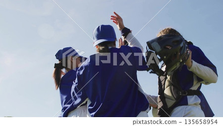 High-fiving female baseball players wearing blue jerseys and caps on sunny field, with catcher gear High-fiving female baseball players wearing blue jerseys and caps on sunny field, with catcher gear 135658954