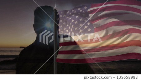 Silhouetted man holding waving American flag on shore at sunset, with sunburst and ocean waves 135658988