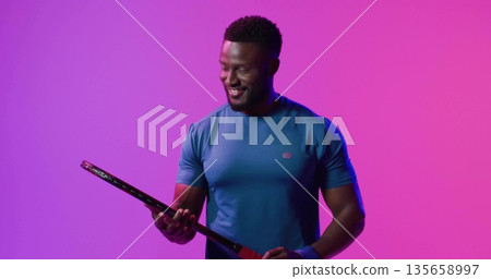 Smiling man wearing blue tee and wristband inspecting black racket under magenta lights in studio 135658997