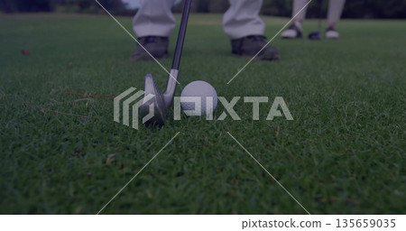 Positioning steel golf iron behind white golf ball on putting green, with golf shoes over grass 135659035
