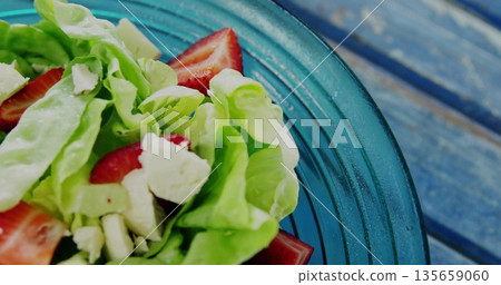 Mixing butter lettuce, halved strawberries, crumbled cheese in blue glass bowl on table, copy space Mixing butter lettuce, halved strawberries, crumbled cheese in blue glass bowl on table, copy space 135659060