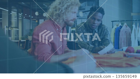 Two designers leaning over table selecting fabric swatches in design studio, with garment samples 135659149