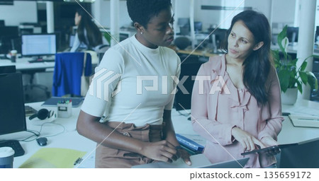 Collaborating women in business attire discussing project at office desk, with laptop and tablet Collaborating women in business attire discussing project at office desk, with laptop and tablet 135659172