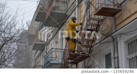 Worker in yellow protective suit climbs metal fire escape on building exterior during winter, with snow-covered branches and balconies visible in the background Worker in yellow protective suit climbs metal fire escape on building exterior during winter, with snow-covered branches and balconies visible in the background 135659298