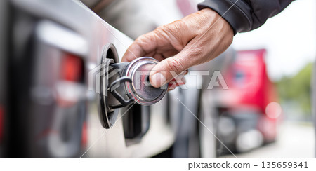 Close-up of a hand operating a fuel cap on a truck, with a blurred background of a red truck and outdoor environment visible 135659341