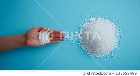 Hand holding small amount of white granules next to a larger pile of granules on a blue background, showcasing size comparison and texture of materials Hand holding small amount of white granules next to a larger pile of granules on a blue background, showcasing size comparison and texture of materials 135659489