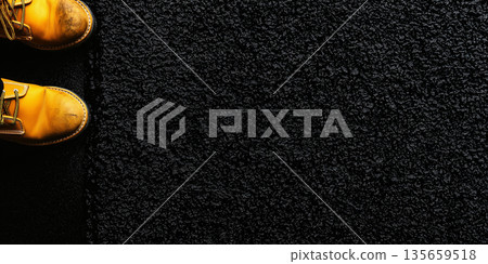 Yellow work boots with scuffed toes positioned on a textured black surface, showcasing the contrast between footwear and ground material in a close-up view 135659518