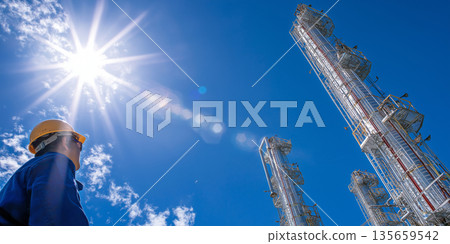 Worker in yellow hard hat gazes up at industrial smokestacks against a bright blue sky with sun rays shining through clouds in a manufacturing facility setting Worker in yellow hard hat gazes up at industrial smokestacks against a bright blue sky with sun rays shining through clouds in a manufacturing facility setting 135659542