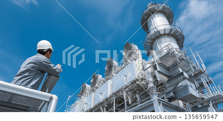Male engineer in gray workwear observes industrial gas turbine equipment against a clear blue sky at a power generation facility Male engineer in gray workwear observes industrial gas turbine equipment against a clear blue sky at a power generation facility 135659547