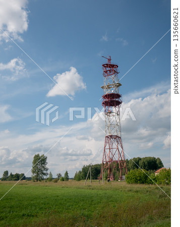 Red and white telecommunication tower in a rural landscape under blue sky. Signal transmission structure for mobile operators 135660621