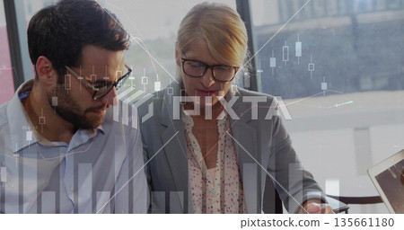 Leaning professionals in blazers reviewing market charts on laptop at desk by window, chart overlay 135661180