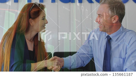 Shaking colleagues wearing business attire sealing financial deal at office desk with chart overlay 135661187