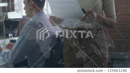 Collaborating analyst and manager reviewing reports on monitor at office, with world map overlay 135661290
