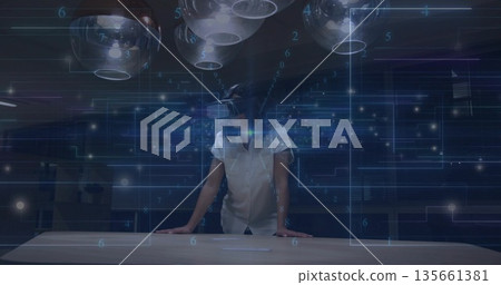 Leaning adult woman wearing VR headset using holographic UI in tech lab, with pendant lights 135661381