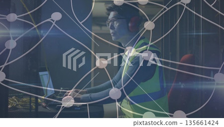 Technician wearing vest, headphones using laptop at data center, with server racks, network cables Technician wearing vest, headphones using laptop at data center, with server racks, network cables 135661424