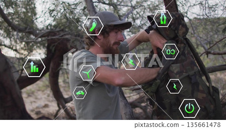 Male hiker fastening camouflage backpack onto branch in dry woodland, with data overlay icons Male hiker fastening camouflage backpack onto branch in dry woodland, with data overlay icons 135661478