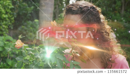 Sniffing woman in pink top and earbud leaning toward peach rose in backyard garden, with flare 135661572