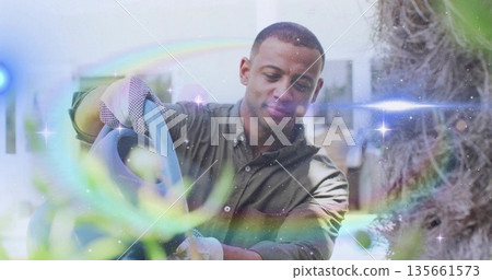 Watering mid adult man nurturing leafy plants in greenhouse holding watering can and wearing gloves 135661573
